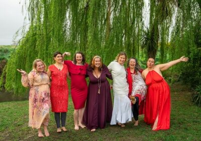 Seven women dressed in red, linking arms and being goofy in front of a willow tree.