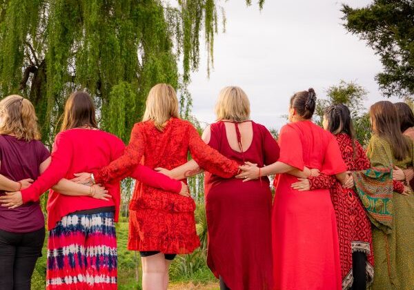 Red Tent Aotearoa Group Hug 2 Women dressed in red, their backs to us, outside under a willow tree, with arms linked.